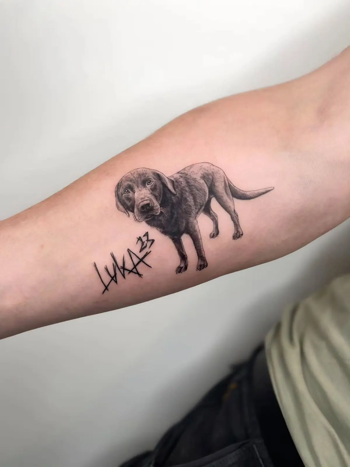 Black and grey portrait of a standing dog with its name lettered underneath, on a forearm.
