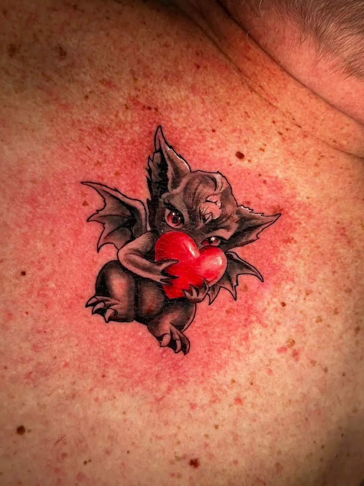 Freshly tattooed cartoon gargoyle with bat wings hugging a glossy red heart, on the chest.