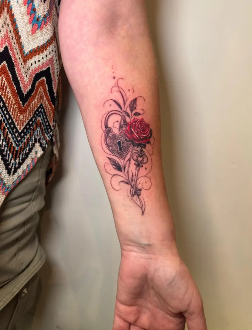 Black and grey heart-shaped padlock and skeleton key joined by filigree, with a small red rose, on an inner forearm.