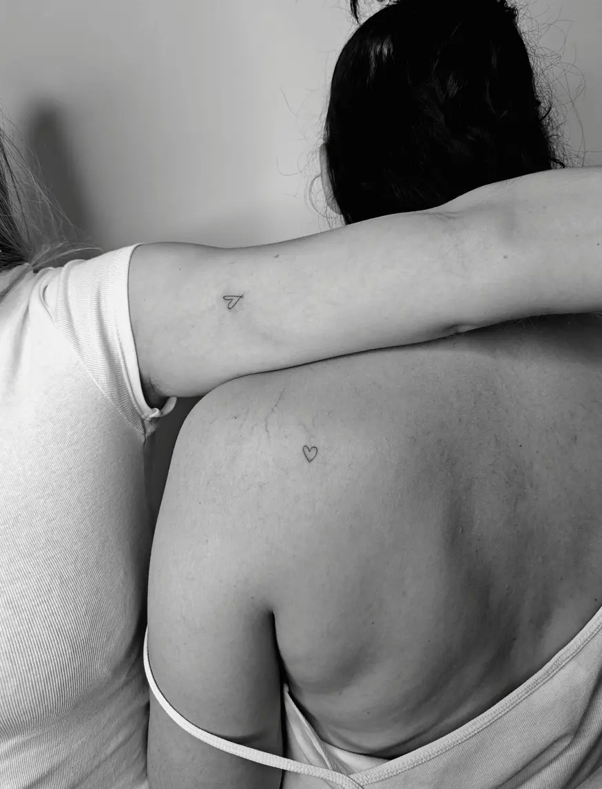 Two friends embracing, one with a tiny paper plane on the arm and the other with a tiny heart on the shoulder.