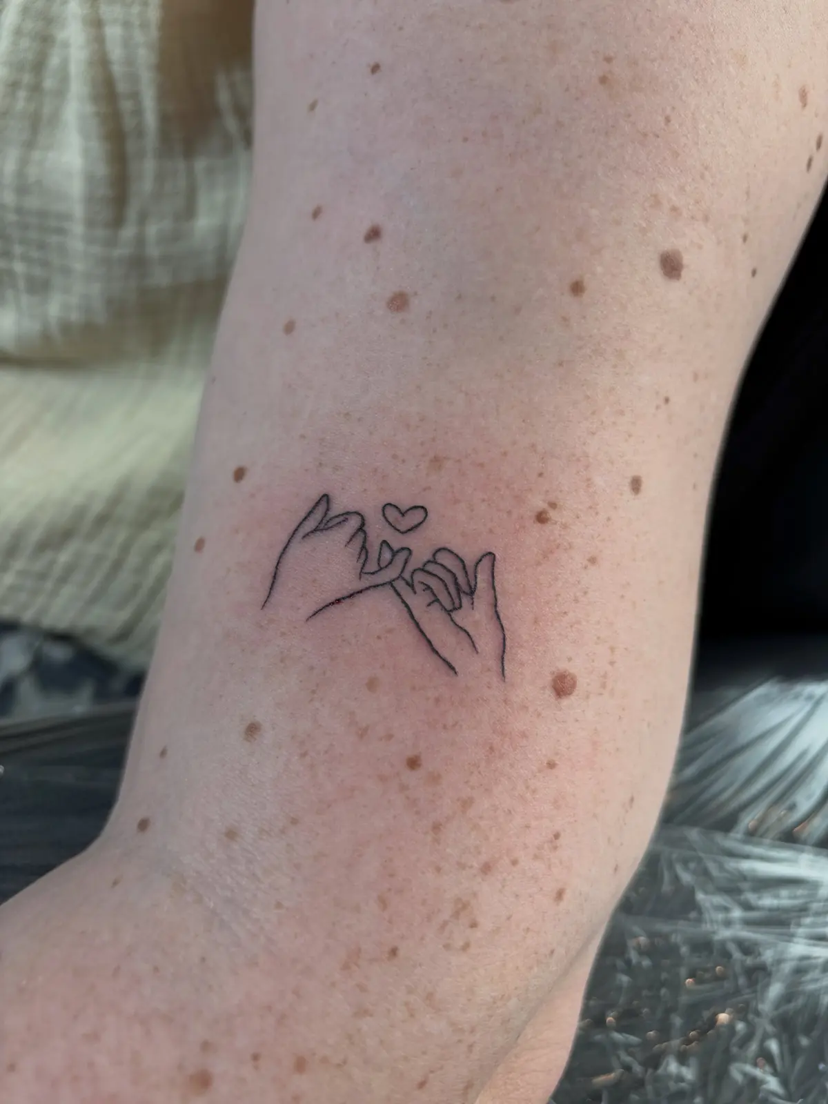 Fine-line drawing of two hands linking little fingers under a tiny heart, on the upper arm.