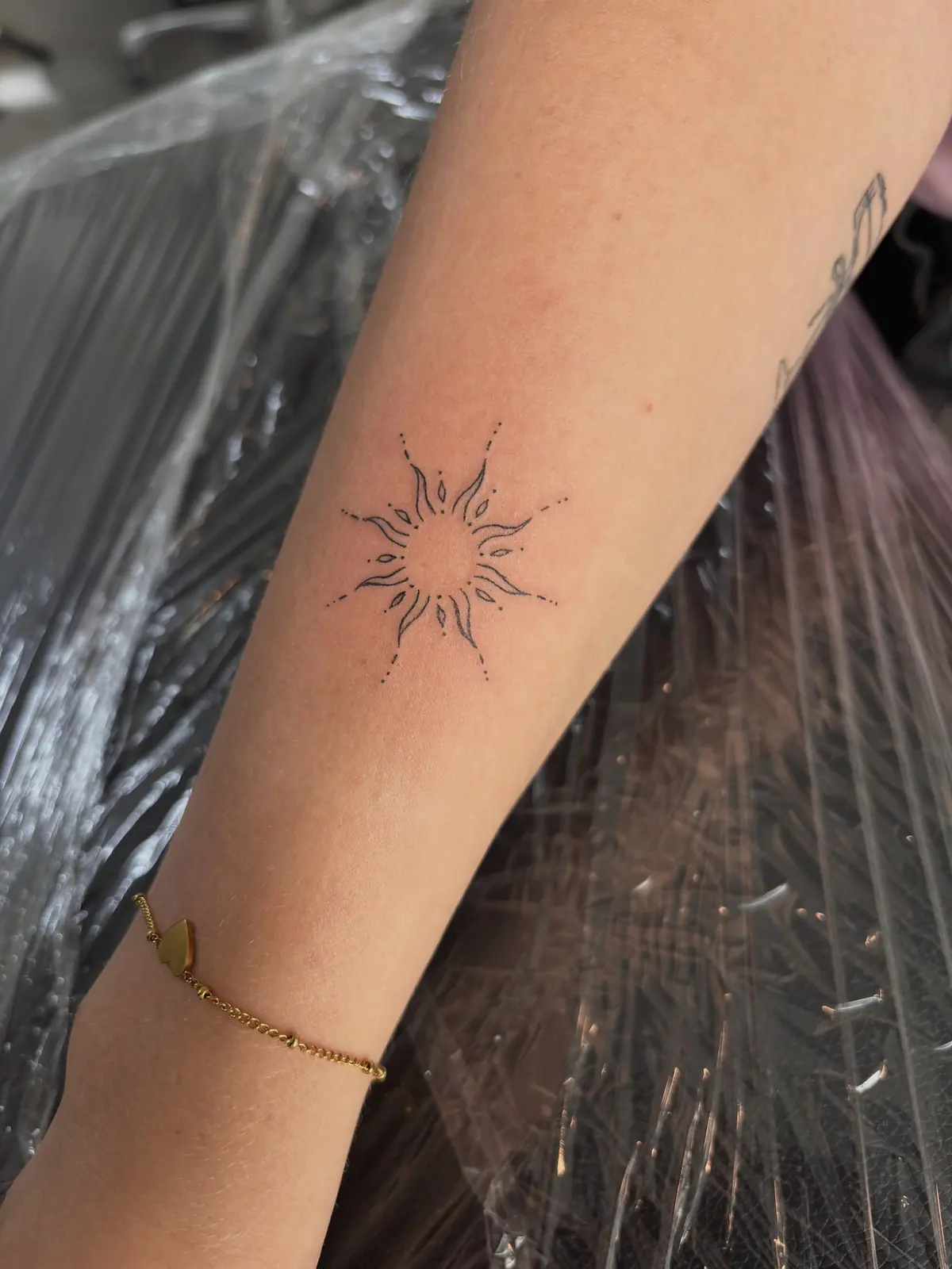 Freshly tattooed fine-line sun with dotted rays on a forearm, resting on a plastic-covered armrest.